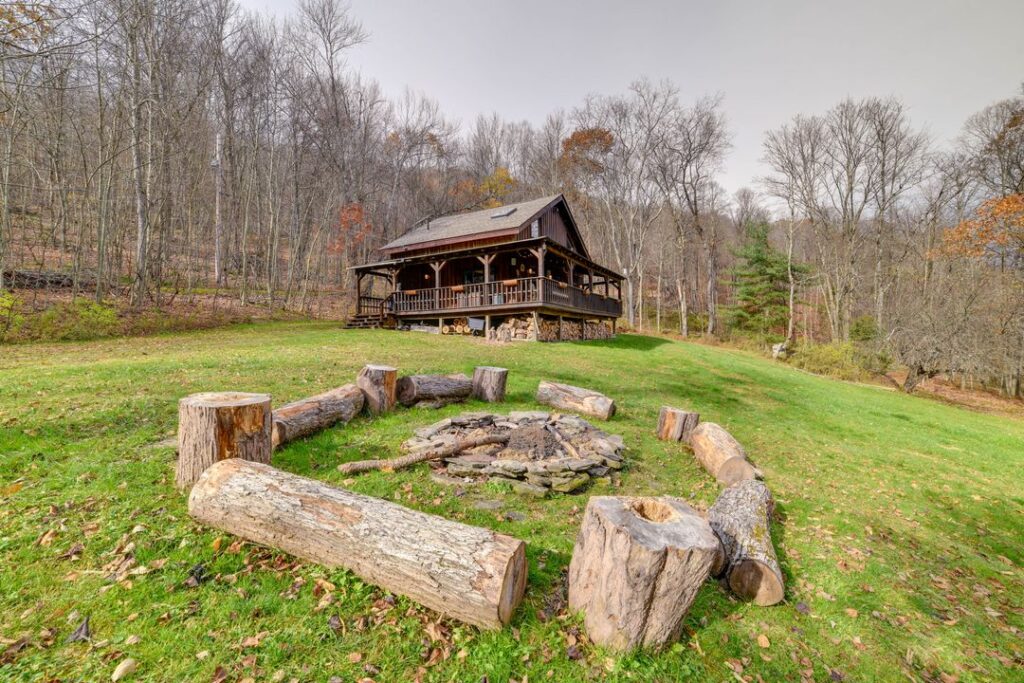 Rustic wooden cabin with a covered wraparound porch, nestled on a grassy hillside surrounded by leafless trees. In the foreground, a circular fire pit made of stacked stones is encircled by large log seats, creating a cozy outdoor gathering space.